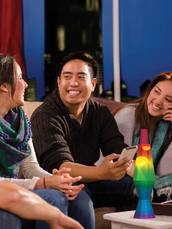 Three friends sit on a couch, chatting and smiling. One holds a phone, while a 14.5" Rainbow LAVA Lamp—perfect for teens and a fun sensory accessory—glows on the table. A cityscape is visible through the window behind them.