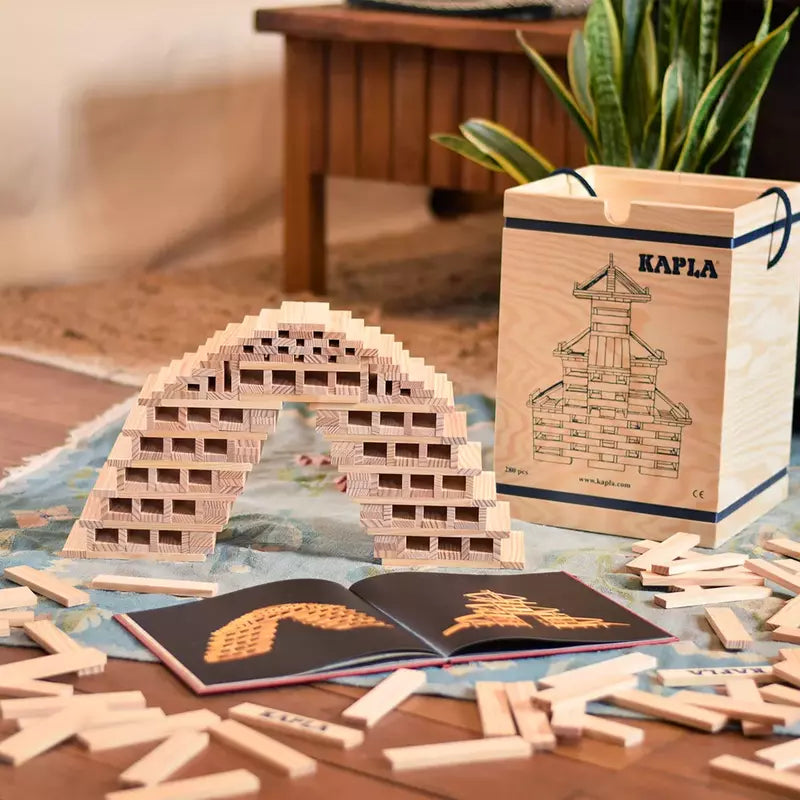 A wooden arch structure crafted from KAPLA® Construction 280 Planks in Wooden Box rests on a rug, showcasing the creativity stimulation of KAPLA's design. Additional planks and an open instruction booklet are strewn about, while the wooden storage box marked "KAPLA" is positioned in the background.