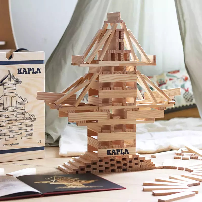 A tower built with KAPLA® Construction 280 Planks in a Wooden Box stands on a surface, with extra planks scattered around to spark creativity. In the background, there's a wooden box labeled "KAPLA" and a book, all set against a softly draped light-colored cloth.