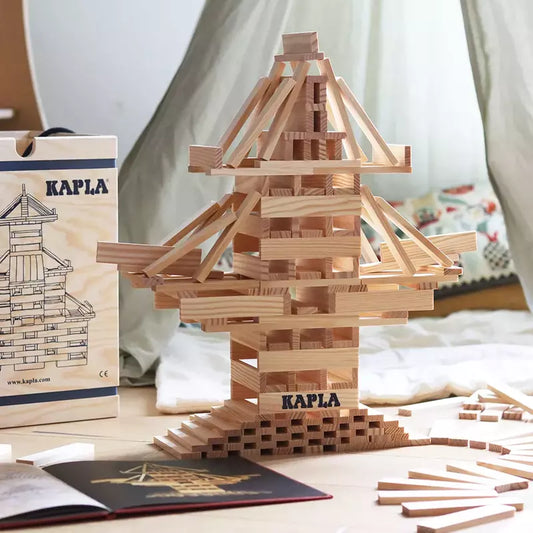 A tower built with KAPLA® Construction 280 Planks in a Wooden Box stands on a surface, with extra planks scattered around to spark creativity. In the background, there's a wooden box labeled "KAPLA" and a book, all set against a softly draped light-colored cloth.