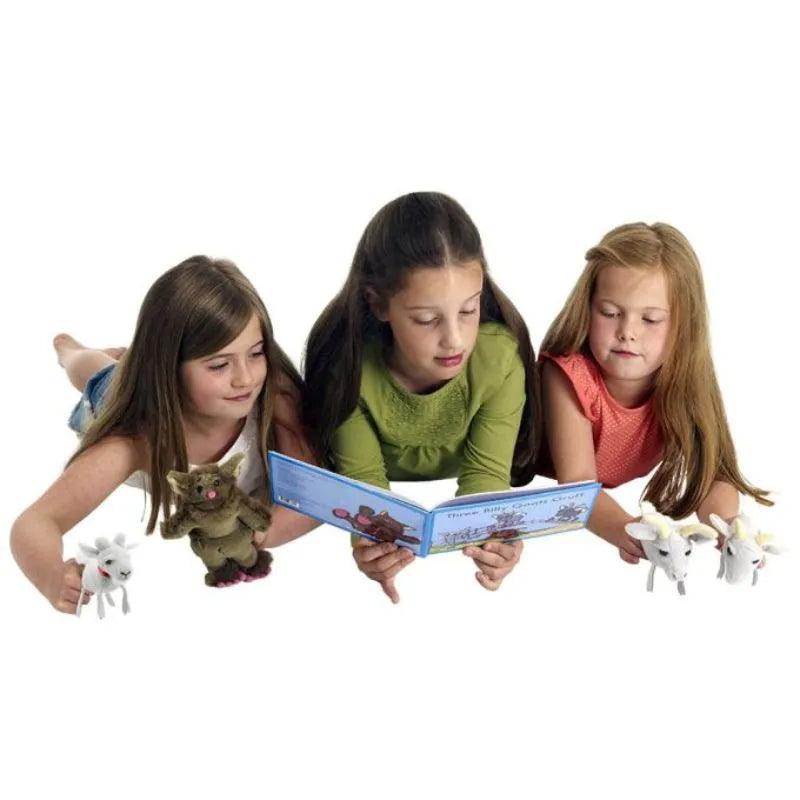Three young girls are lying on their stomachs, reading a book together. They are surrounded by various animal figurines, including a brown monster and a couple of white unicorns from The Puppet Company Finger Puppet Story Set Three Billy Goats Gruff. Among the colorful toys, finger puppets add a touch of magic to their storytelling adventure.