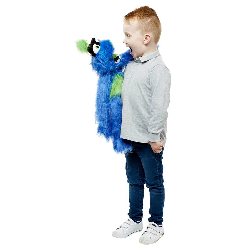 A brown-haired child, dressed in a light gray shirt and dark jeans, joyfully interacts with The Puppet Company Blue Monster on his arm against a white backdrop.