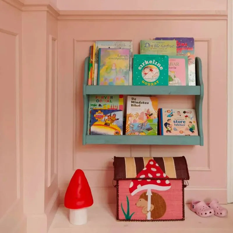 A pastel pink nursery wall features a teal bookshelf with colorful books, the Lamp Small Mushroom Red, a handcrafted lamp, a toy chest with hedgehog and mushroom designs, and pink clogs—a charming touch for children's room decor.