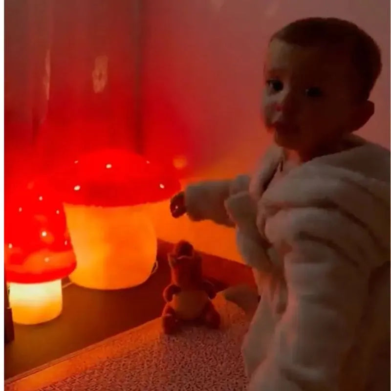 A young child in a fluffy white robe stands near the glowing Lamp Small Mushroom Red and a plush toy, with warm ambient lighting creating a cozy atmosphere—perfect for unique children's room decor.