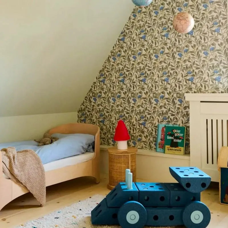 A cozy kids’ bedroom with a small bed, blue pillow, blanket, soft toy, floral wallpaper, wicker side table featuring the Lamp Small Mushroom Red, children’s books, and a blue toy car—perfect for charming children’s room decor.
