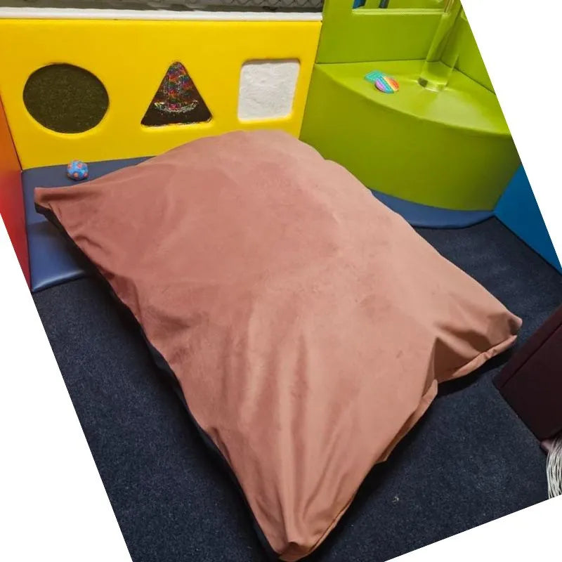 A Sensory Crash Bag Large, tan in color, sits on a dark carpet in a vibrant play area with padded yellow and green panel walls featuring triangle, circle, and square shapes. A small toy nearby adds extra therapeutic benefits.