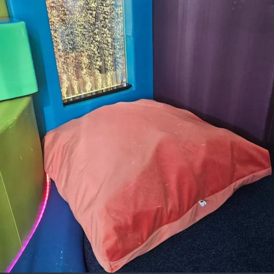 A small, orange Sensory Crash Bag sits on a dark carpeted floor in a colorful room with soft foam blocks and a sensory bubble tube in the background, providing therapeutic support for emotional regulation.
