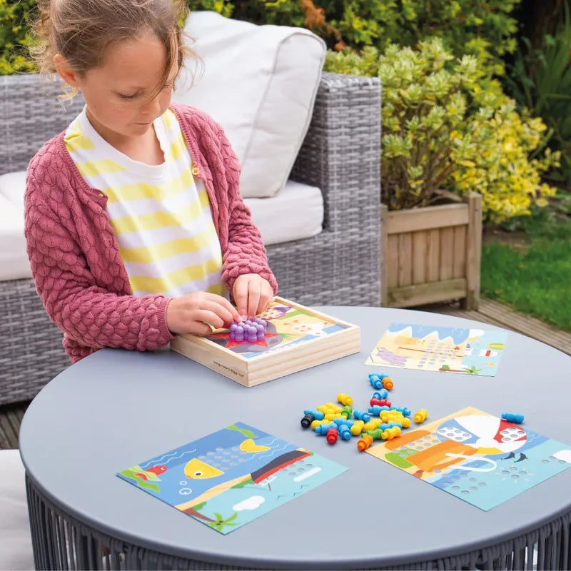 A child in a pink cardigan plays with the Bigjigs Seaside Peg Board on a round gray table, boosting fine motor skills. The table is dotted with colorful game pieces and illustrated cards, set on an outdoor patio adorned with wicker furniture and lush greenery in the background.