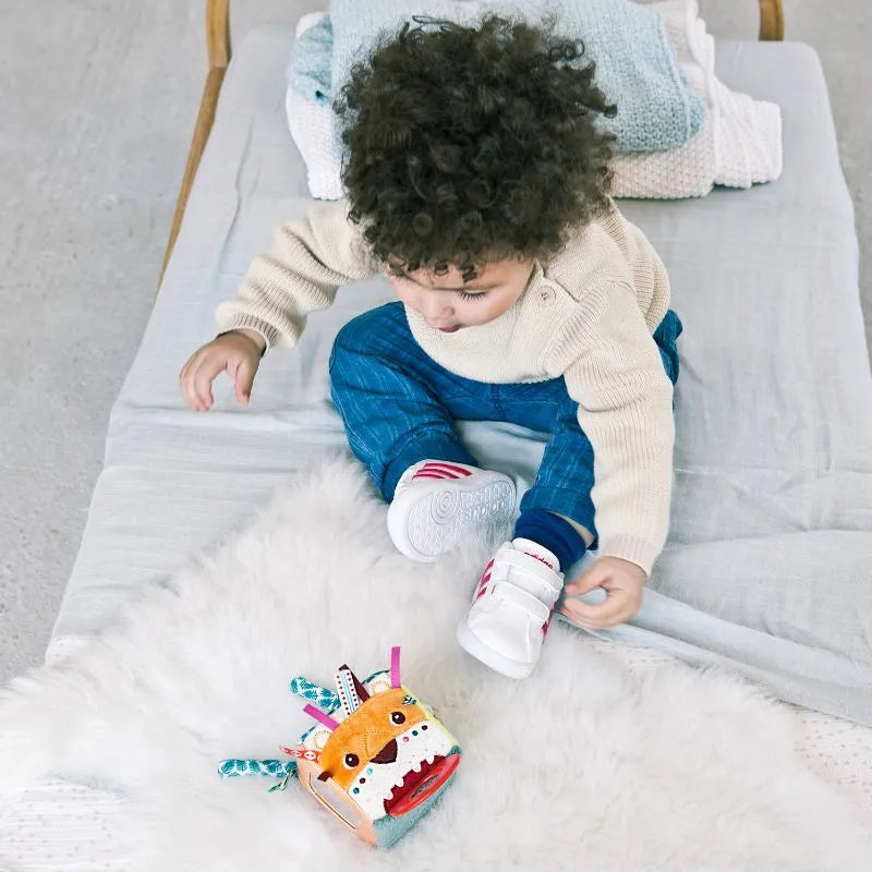 A toddler with curly hair sits on a bed, wearing a beige sweater, blue pants, and white sneakers. The child explores developmental stages while gazing at a colorful Lilliputiens I'm Discovering Jack Activity Cube and teething ring on the fluffy white rug.