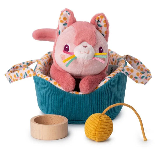 The Lilliputiens I'm Exploring Jeanne Cat With Basket features a plush pink cat toy with rainbow whiskers, nestled in a teal floral-handled basket. It includes a small wooden bowl and textured yellow ball on a string, perfect for nurturing caring skills as a children's toy.