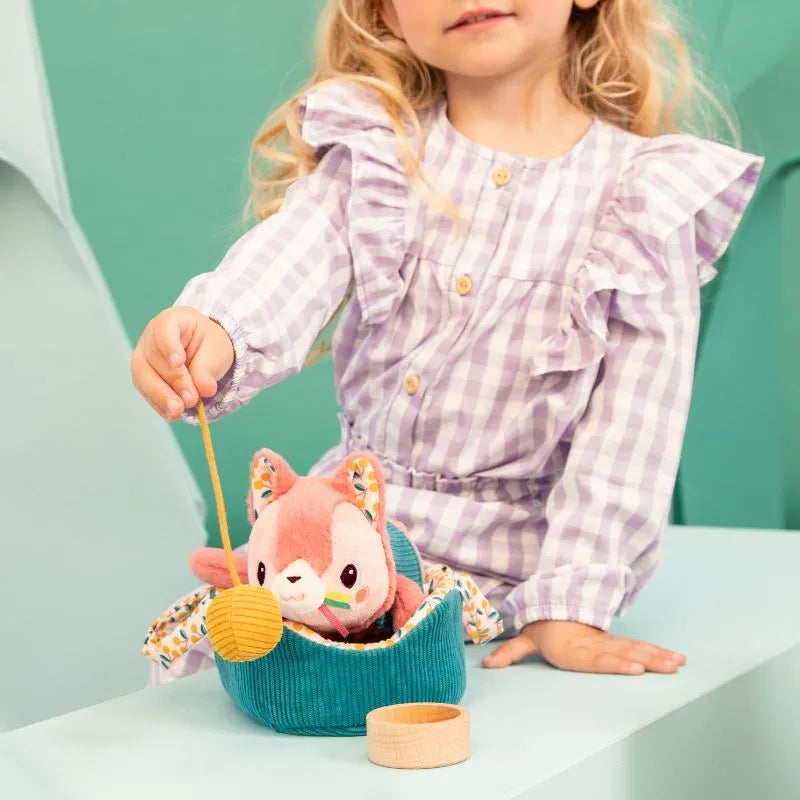 A child in a checkered purple dress cares for the Lilliputiens I'm Exploring Jeanne Cat With Basket, featuring a plushie fox in a soft blue basket with a small yellow bag. This toy encourages nurturing skills against a backdrop of soft green.