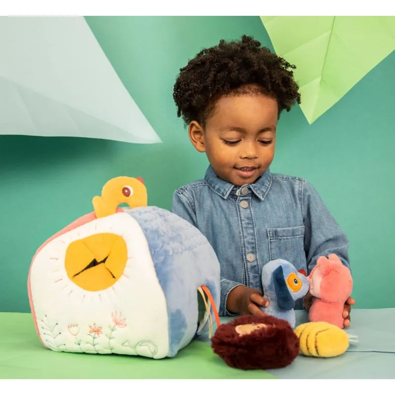 A child with curly hair, in a denim shirt, plays with the Lilliputiens I'm Discovering Jules' House And 4 Hidden Rattles alongside soft toy animals. A light green background with geometric shapes makes it perfect for peek-a-boo games and endless fun.