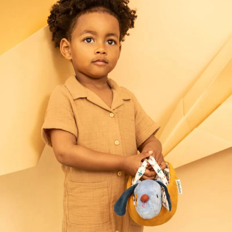 A young child in a light brown romper stands against a beige geometric background, holding the Lilliputiens I'm Exploring Jules Dog With Basket toy. The curly-haired child, gazing slightly off-camera with a neutral expression, resembles a curious puppy.
