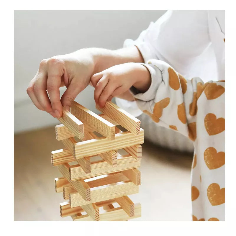 A child wearing a heart-patterned shirt is creatively stacking KAPLA® Construction 200 Planks on a table with the guidance of an adult's hand, in an indoor setting that emphasizes the focus on hands and blocks.