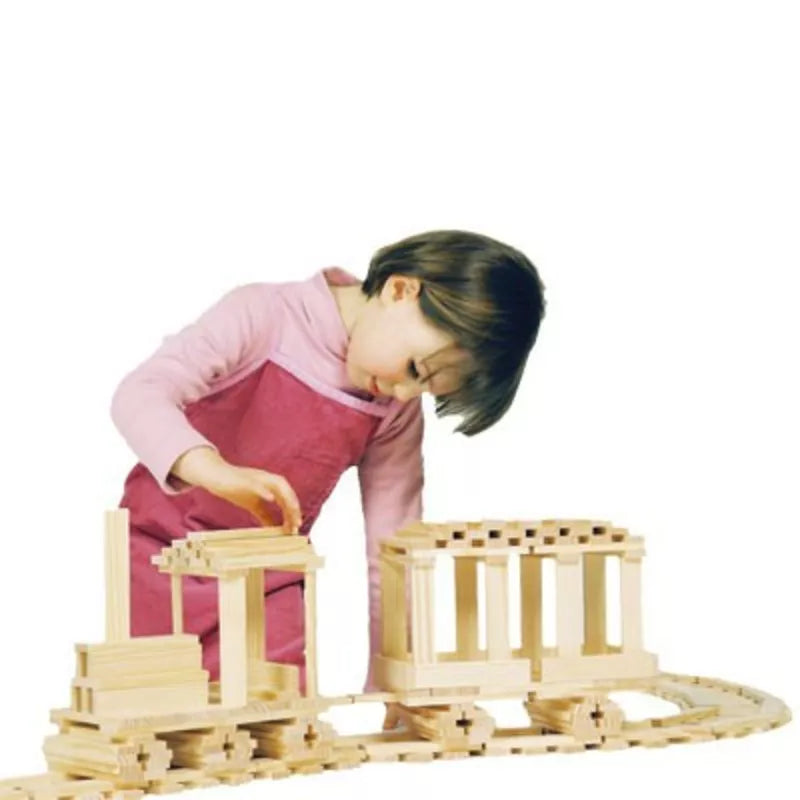 A young child in a pink and red outfit is deeply engaged in building with the KAPLA® Construction 200 Planks. The wooden set, featuring interlocking pieces much like those of a train, captures the child's creative focus as they determinedly add a new piece against the white background.