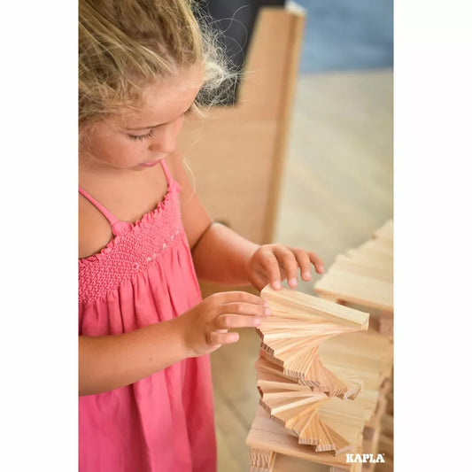 A young girl in a pink dress meticulously constructs an imaginative tower using KAPLA® Construction 200 Planks.