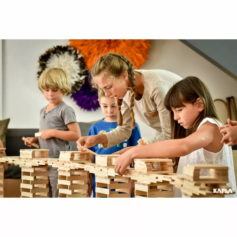 Four children are focused on building structures with KAPLA® Construction 200 Planks in a room adorned with colorful wall decorations. They're collaborating and concentrating on their constructions, unleashing creativity at every turn.