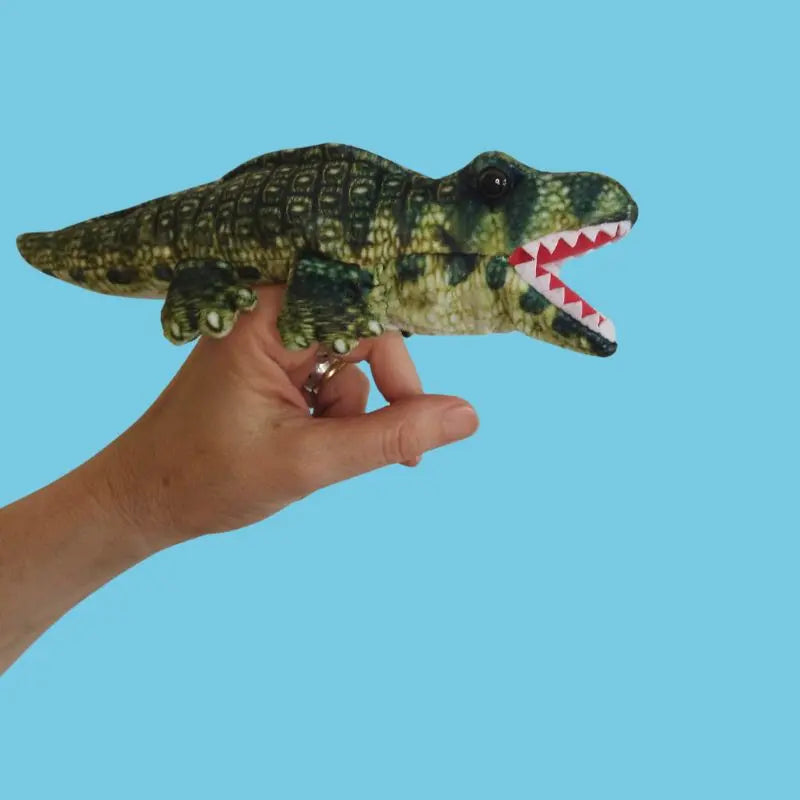 A hand holds The Puppet Company Alligator Finger Puppet from the Little Creatures Range, showing its open green mouth and white teeth against a plain blue background.
