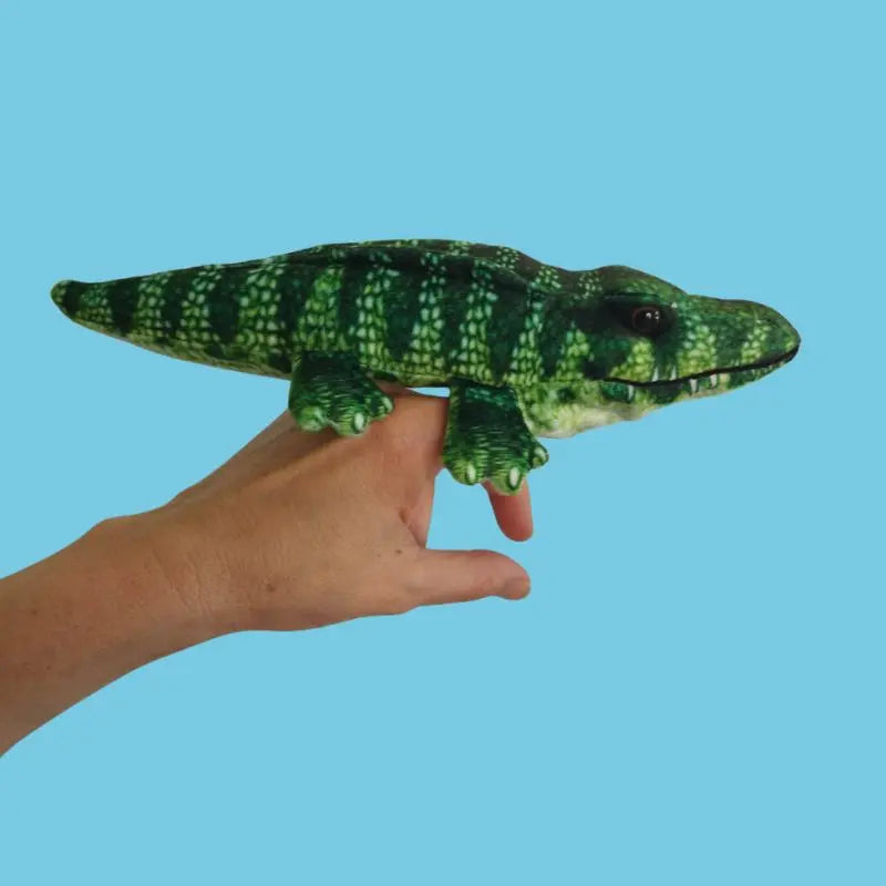 A hand holds The Puppet Company Crocodile Finger Puppet – Little Creatures Range, its green and yellow colors ready for playful adventures against a plain blue background.