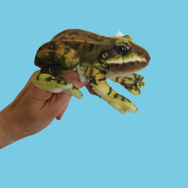 A hand holds The Puppet Company Green Frog Finger Puppet from the Little Creatures Range against a solid light blue background—perfect for storytime fun.