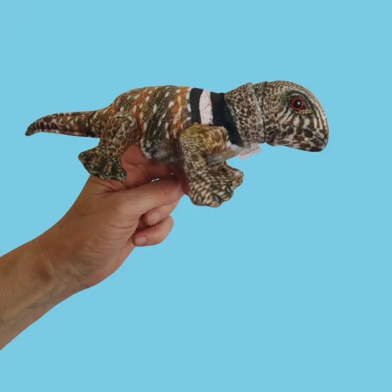 A hand holds The Puppet Company Lizard Finger Puppet from the Little Creatures range, featuring textured, multicolored fabric—perfect for nature-loving kids—against a plain light blue background.