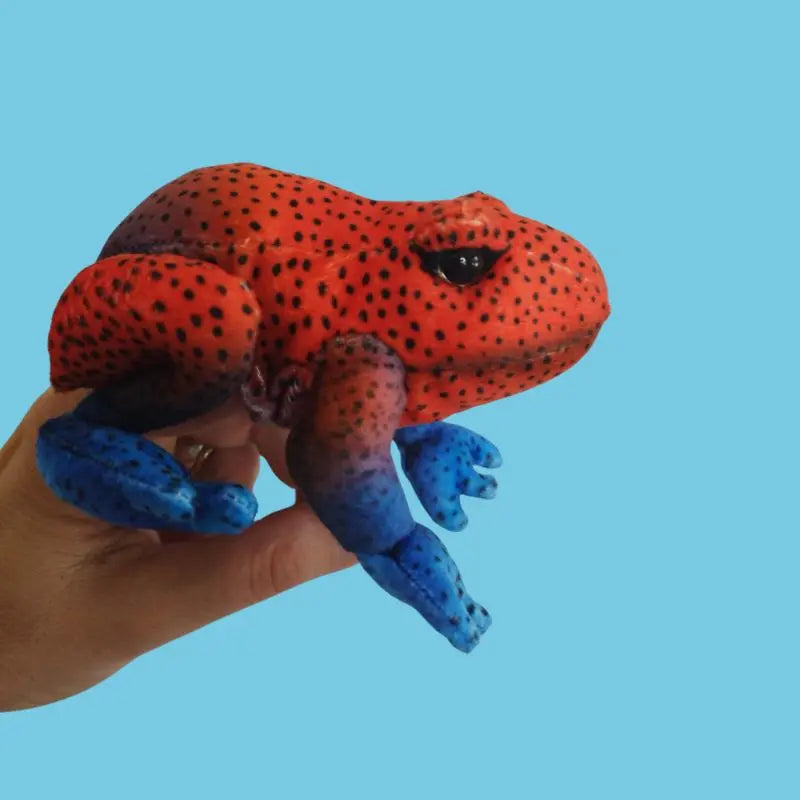 A hand holds The Puppet Company Red Frog Finger Puppet from the Little Creatures Range—a vibrant red frog with black spots and bright blue, spotted legs—against a plain light blue background.