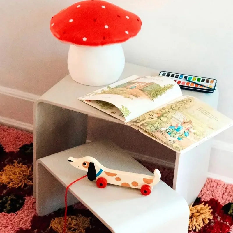 A cozy children’s reading nook features a Lamp Large Mushroom Red, an open picture book, paint box, and wooden pull-toy dog on a small white table with a colorful carpet—charming children's bedroom decor handcrafted in Germany.