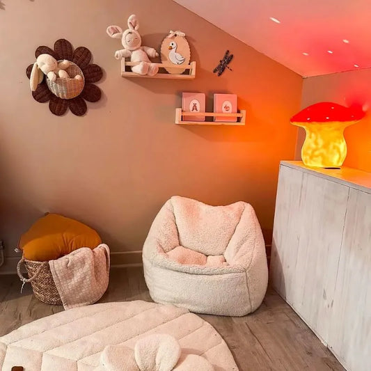 A cozy children's room features a soft armchair, round cushioned rug, plush animal toys on shelves, and the Lamp Large Mushroom Red glowing on a white cabinet under warm lighting—perfect for charming kids' decor.