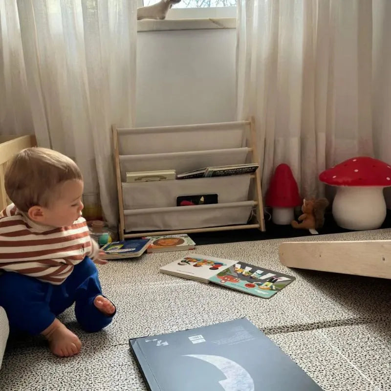 A baby sits on the floor of a cozy children's bedroom, surrounded by picture books, a wooden bookshelf, and the Lamp Large Mushroom Red. Soft light from the lamp filters through sheer curtains in the background.
