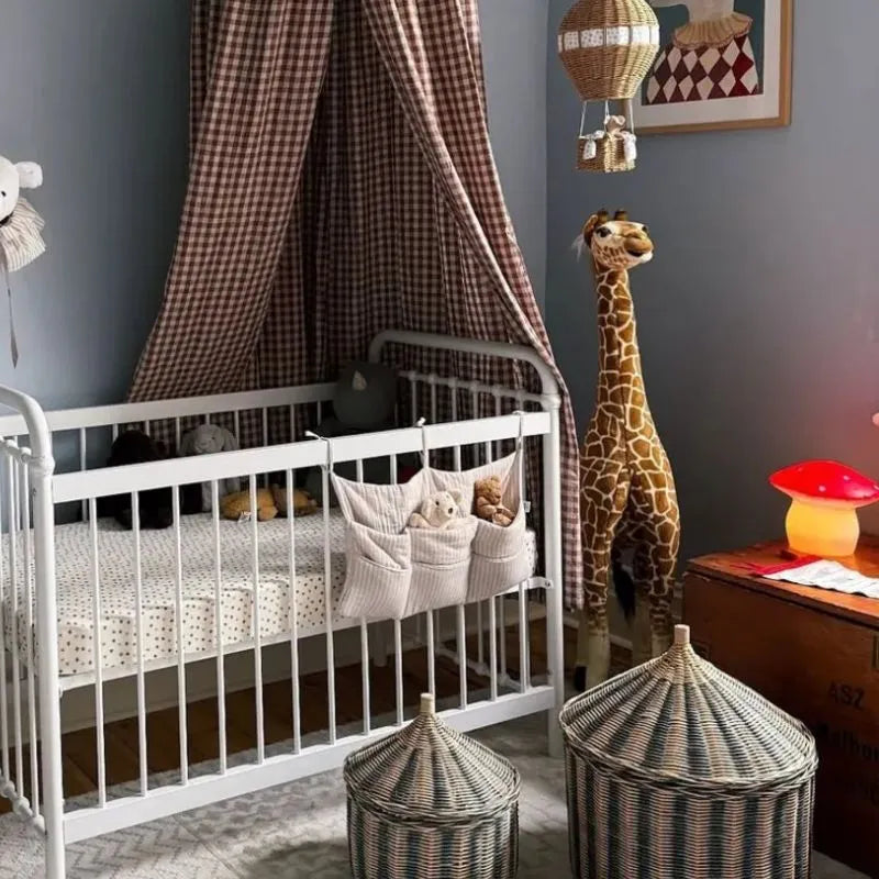 Cozy nursery featuring a white crib, stuffed animals, and a tall plush giraffe. A brown gingham canopy hangs above. Two woven baskets and the Lamp Medium Mushroom Red are near a wooden chest beneath soft lighting.