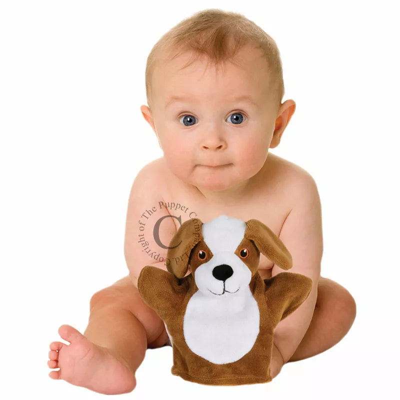 A baby sits against a white backdrop, holding "The Puppet Company My First Puppet Dog" in brown and white. With big eyes and a slight smile, the baby engages in interactive play, one leg bent and the other outstretched, fostering early language development through imaginative interaction.