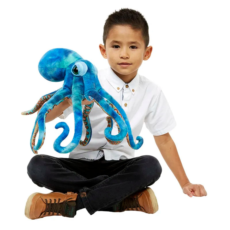 A child wearing a white shirt and black pants sits cross-legged while holding The Puppet Company Large Creature Octopus, ideal for imaginative play. The child looks directly at the camera with a neutral expression.