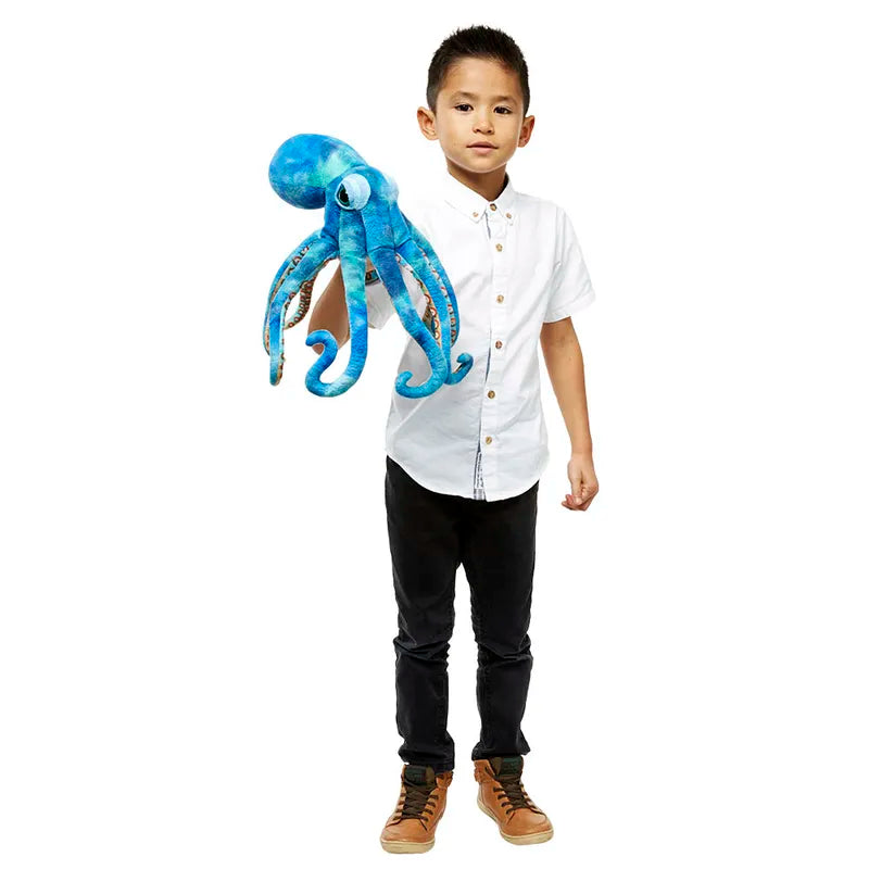 A child in a white shirt and black pants holds The Puppet Company Large Creature Octopus, a large blue plush toy ideal for imaginative play, against a plain white background.