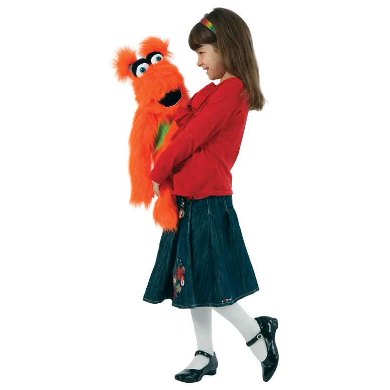 A young girl in a red sweater and dark skirt holds The Puppet Company Orange Monster, featuring bright fuzzy fur and googly eyes. Smiling warmly at the puppet, she is framed against a plain white background.