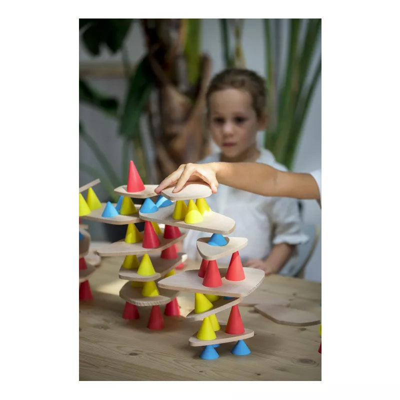 A child enhances creativity and development by building a colorful structure with the Piks Construction Big Kit – 64 pieces, using wooden discs and red, yellow, blue cones on a table. The background is slightly out of focus.