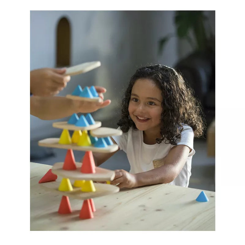 A curly-haired child cheerfully plays with the Piks Construction Big Kit, a balancing game featuring colorful wooden pieces. Assisted by another person adding a piece, they sit at a wooden table in a well-lit room, nurturing children's development through engaging play.
