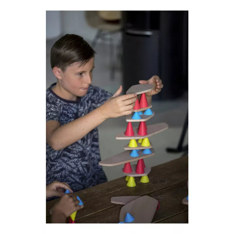A child in a blue patterned shirt creatively focuses on building with the Piks Construction Big Kit – 64 pieces, balancing red, yellow, and blue wooden cones and planks on the table to explore development through playful indoor experimentation.