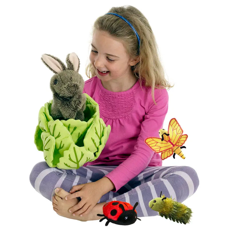 A girl in a pink shirt and striped pants sits cross-legged, smiling at The Puppet Company's Hide-Away Rabbit nestled in a fabric cabbage. Around her, plush toys of a ladybug, caterpillar, and yellow butterfly create a whimsical scene.