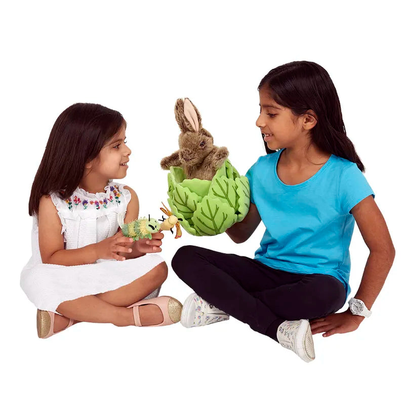 Two young girls sit cross-legged on a white background, smiling. One holds "The Puppet Company Hide-Away Rabbit" glove puppet, its large ears partially hidden in green leaves, while the other holds a stuffed bird.