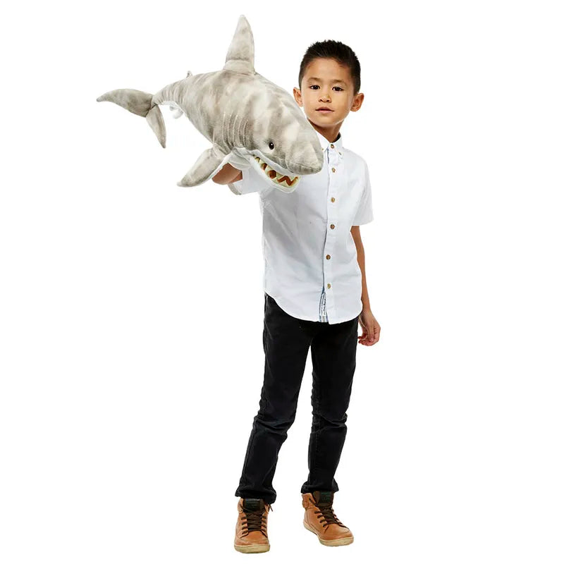 A young boy stands against a plain white background, wearing a white shirt and black pants, holding The Puppet Company Large Creature Shark with its mouth open. Immersed in imaginative play, he brings the natural world to life.