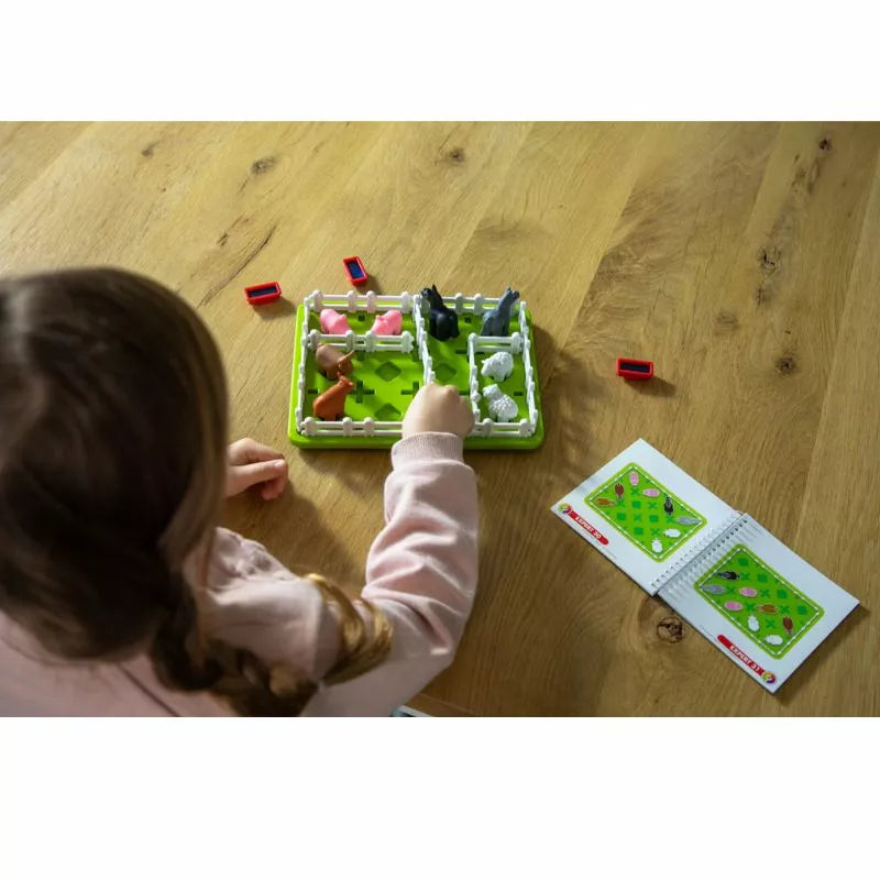 A child is captivated by the SmartGames Smart Farmer, arranging animal figures on a vibrant green board at a wooden table. The open booklet guides them in solving puzzles through specific figure patterns for an engaging playtime.