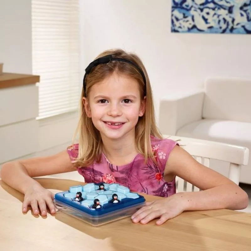 A blonde girl in a pink floral dress smiles while playing SmartGames Penguins Pool Party, a penguin-themed puzzle game, at a table in a brightly lit, minimally decorated room.