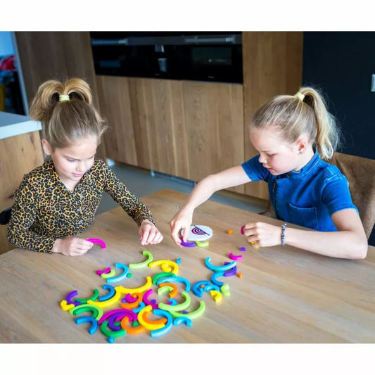 Two children with ponytails and colorful bows sit at a wooden table, deeply engaged in the Buitenspeel Speed Bows Game. They focus on assembling the vibrant, curved puzzle pieces amidst a modern kitchen backdrop.