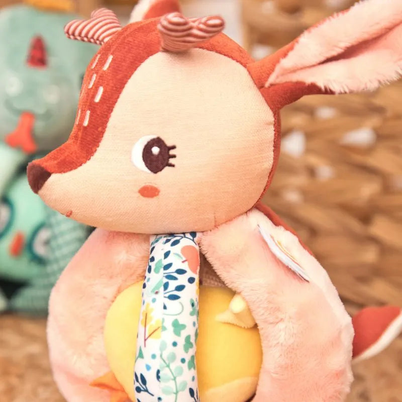 The Lilliputiens I'm Discovering Stella The Faon Rattle With Handles is a soft, brown and cream plush deer toy with large stitched eyes and floral fabric on its neck. It holds a yellow textured baby rattle ball. Another plush in blue and green hues can be seen partially in the background.