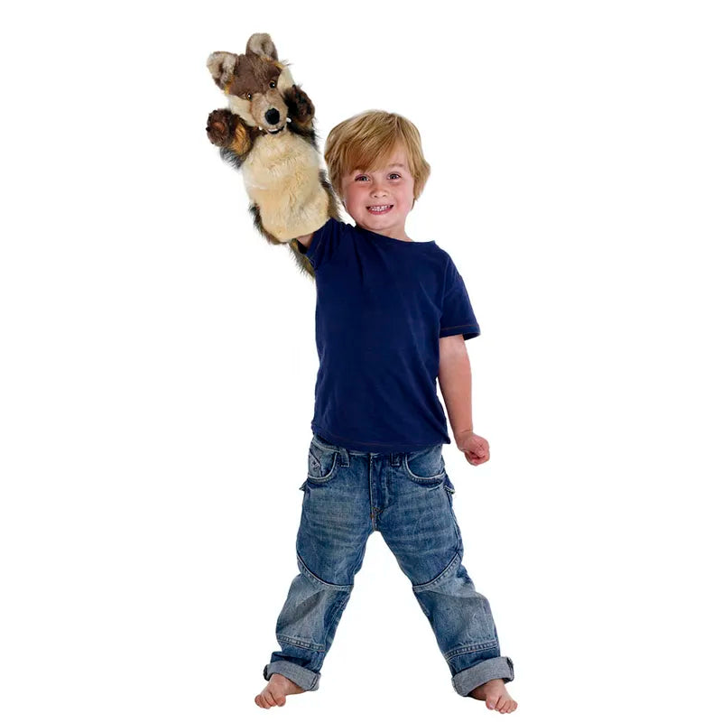A joyful, barefoot young child with blonde hair in a navy blue shirt and jeans holds up The Puppet Company Long Sleeved Puppet Wolf against a plain white background, smiling brightly and ready for creative puppet shows.