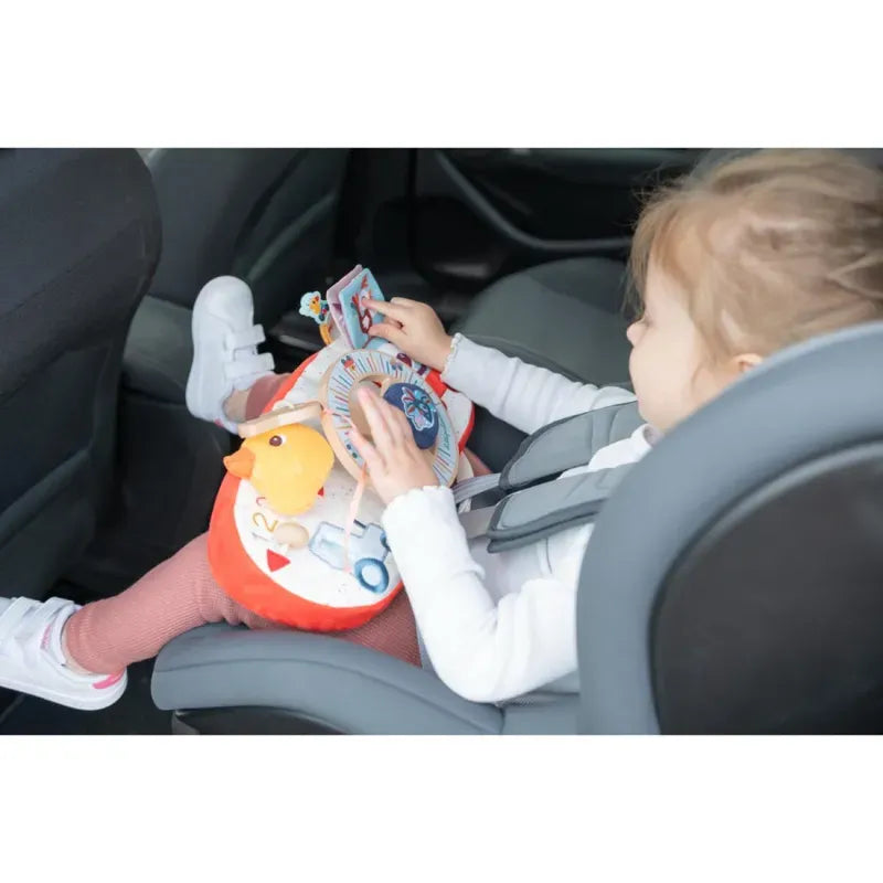 A young child sitting in a car seat, engaged with the colorful Lilliputiens Farm Car Activity Panel on their lap. The baby toy features a variety of items including a small mirror, plush objects, and textured elements. The child, comfortably outfitted in a white long-sleeve shirt and pink pants, enjoys car journeys with this entertaining companion.