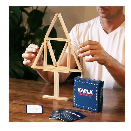 A person in a white shirt assembles a wooden geometric structure using planks. Nearby, the "Kapla Challenge" cards and box hint at mastering more than just building—it's about conquering the balance game.