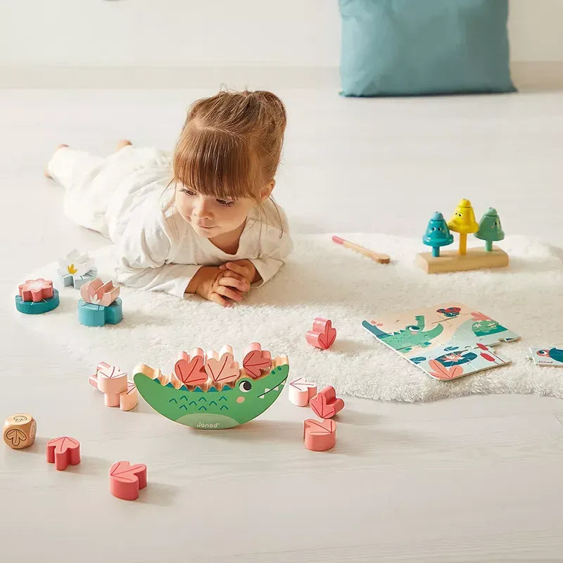 A young girl lies on a white rug, playing with colorful wooden toys. She is focused on a Janod Box of Toys 24 Months, one of many developmental toys around her. Other sensory and educational toys, including a wooden tree set and a puzzle, are scattered on the rug. A blue cushion rests in the background.