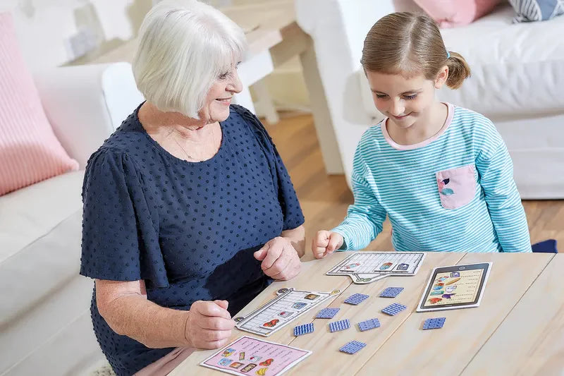 An elderly woman and a young girl are happily engaged in playing Orchard Toys Shopping List together at a table. They interact with the game pieces with smiles and concentration. The room exudes a cozy and bright atmosphere, featuring a white couch adorned with pink cushions in the background.