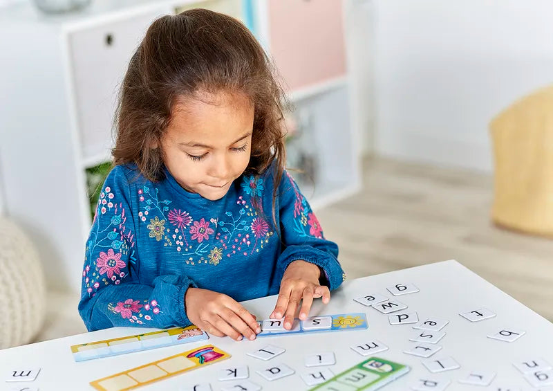 A young girl with long hair, wearing a blue floral dress, sits at a table arranging the Orchard Toys Match And Spell tiles. She appears focused on creating words. The room is bright and decorated in soft colors.